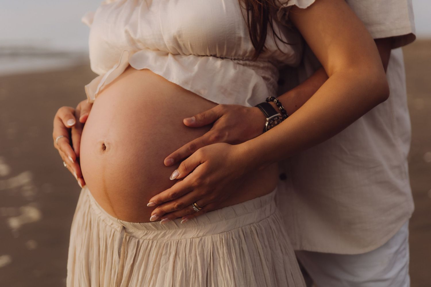 Pregnant woman and partner cradling baby bump together on beach at sunset.