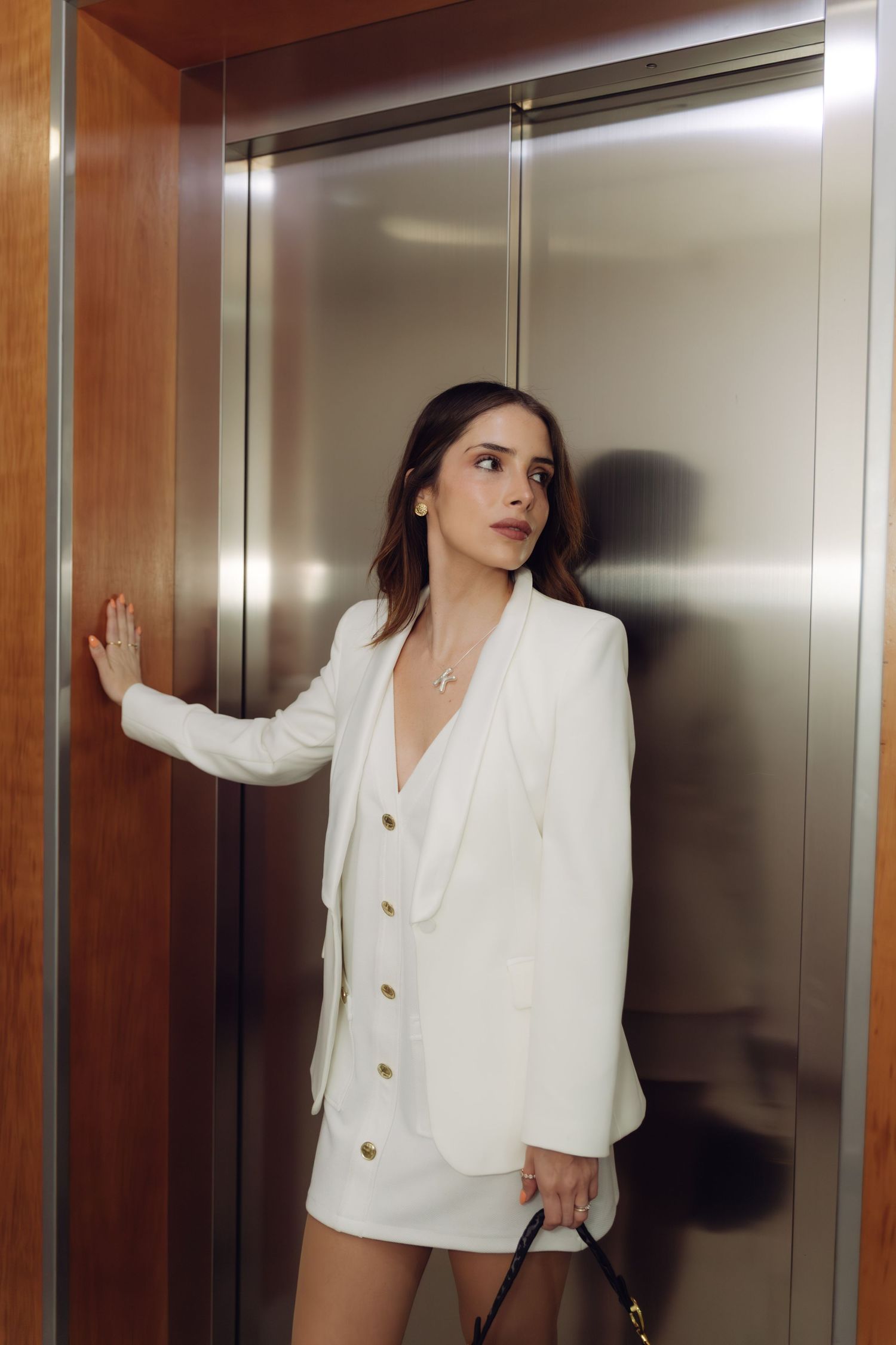 Stylish woman in white blazer and mini skirt holds handbag while standing in modern elevator.