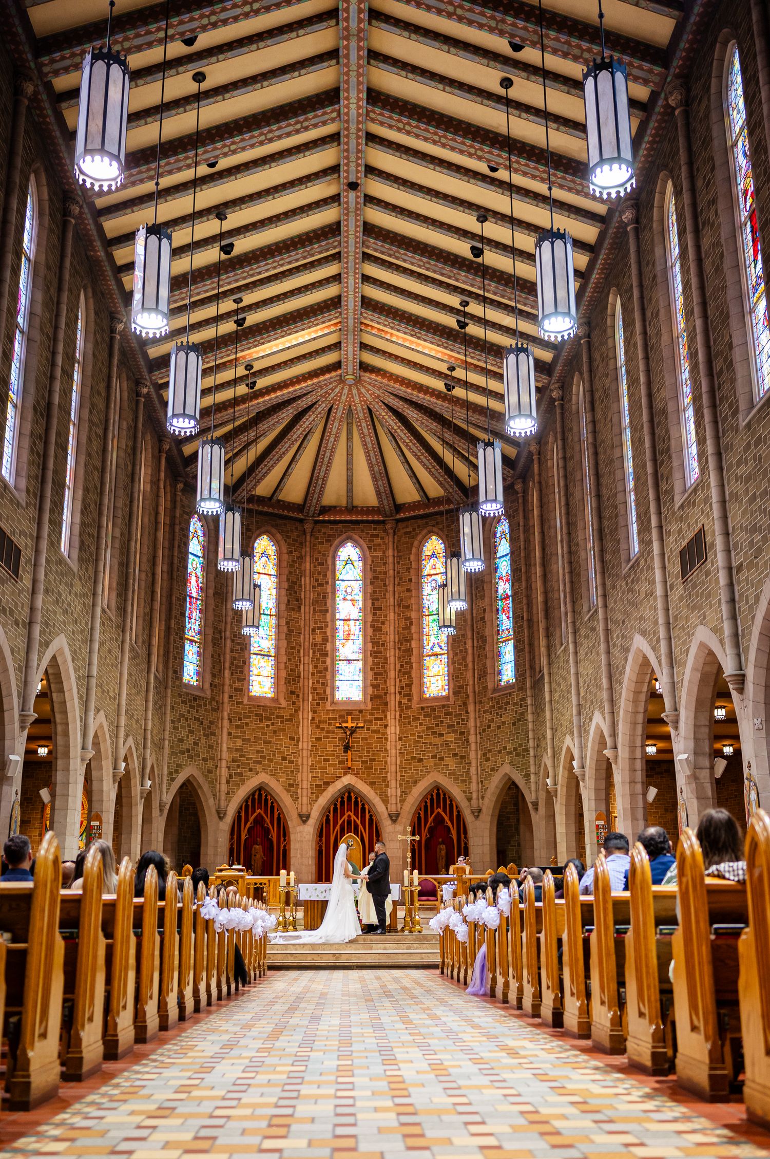 Elegant church wedding ceremony with bride and groom at altar, stained glass windows, wooden pews, and vaulted ceiling.