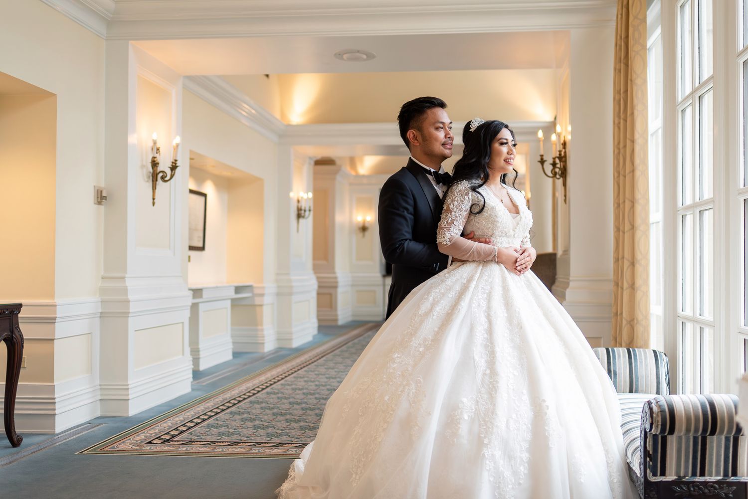 Elegant bride in lace ballgown and groom in tuxedo posing in a grand wedding venue hallway.