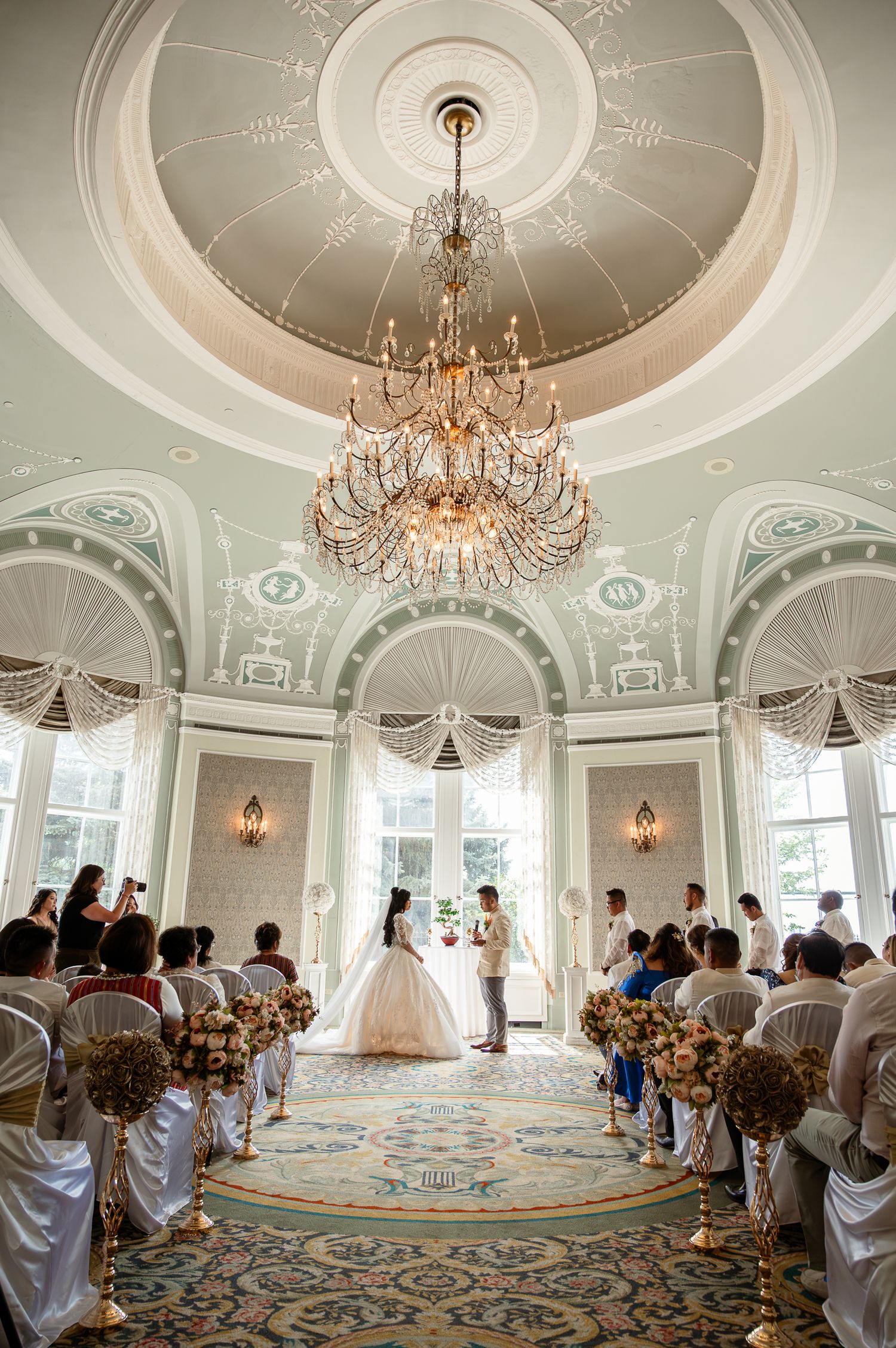 Elegant wedding ceremony in ornate ballroom with crystal chandelier, bride in white gown, guests seated in decorated aisles.