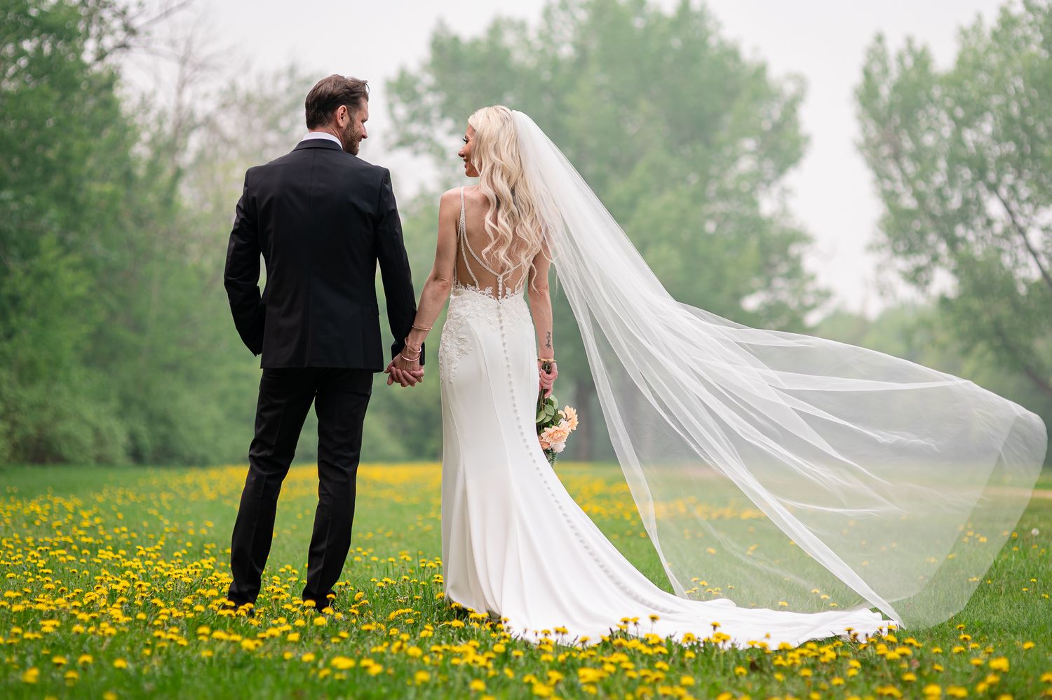 Bride and groom holding hands in a field of yellow wildflowers, bride's veil flowing in the wind.