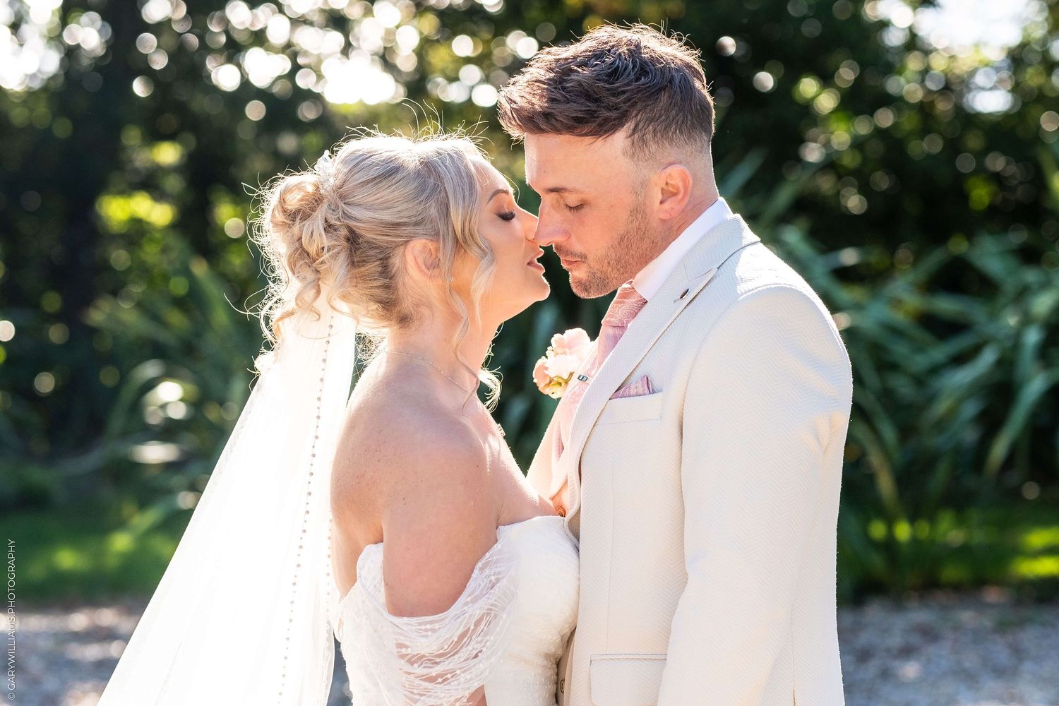 A tender intimate moment between newlyweds in formal wedding attire standing outdoors in natural sunlight.