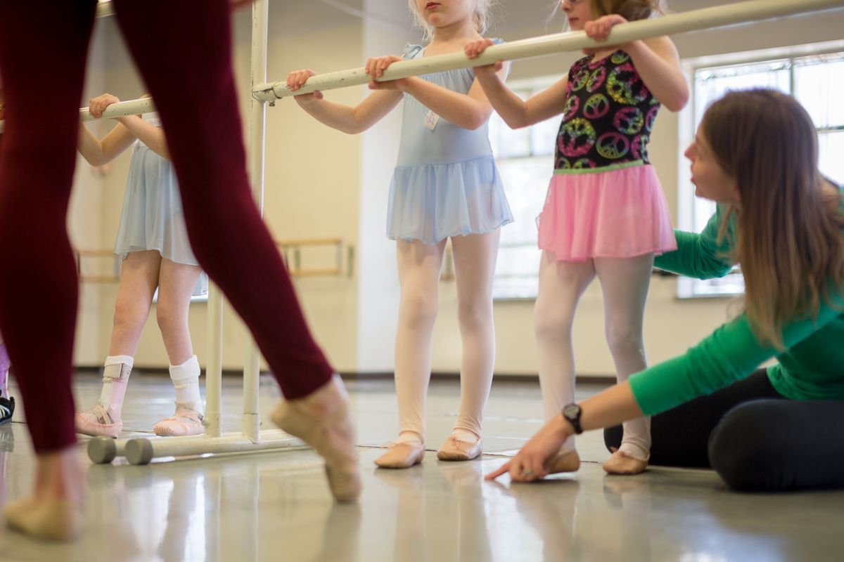 Young ballet students practice at the barre in a dance studio wearing tutus and ballet slippers.