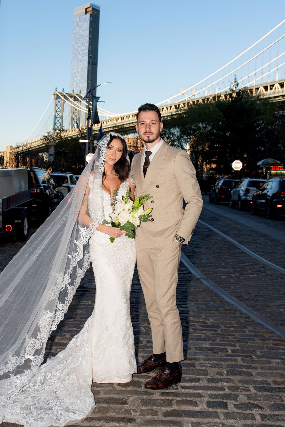 Wedding couple poses on cobblestone street with Manhattan Bridge and urban skyline in background.