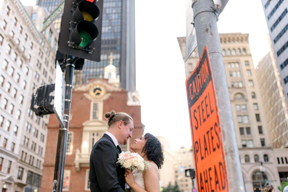 A couple shares a romantic kiss on a busy city street corner with tall buildings and traffic lights in the background.