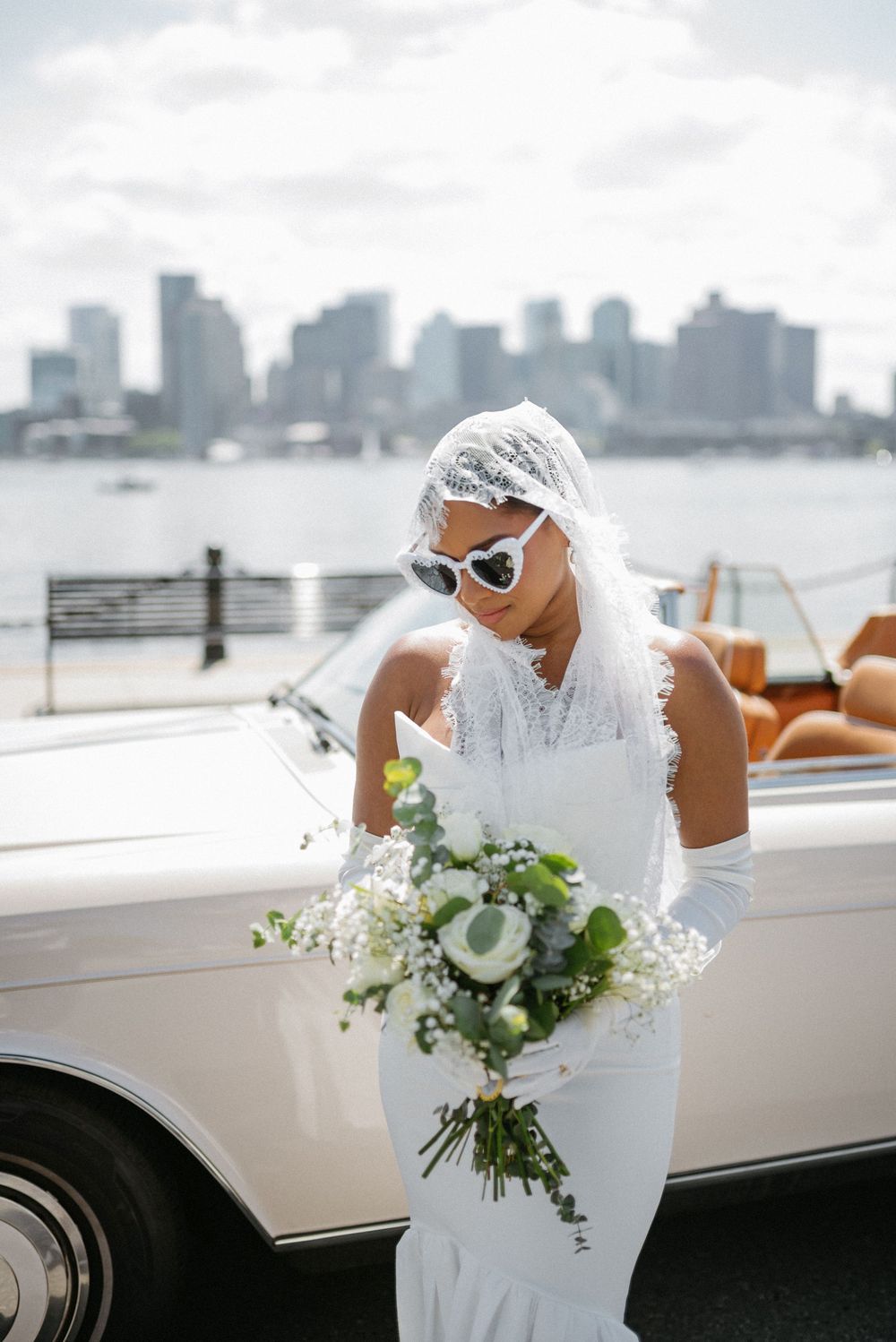 Classic white vintage car parked by waterfront with city skyline and floral bouquet in view.