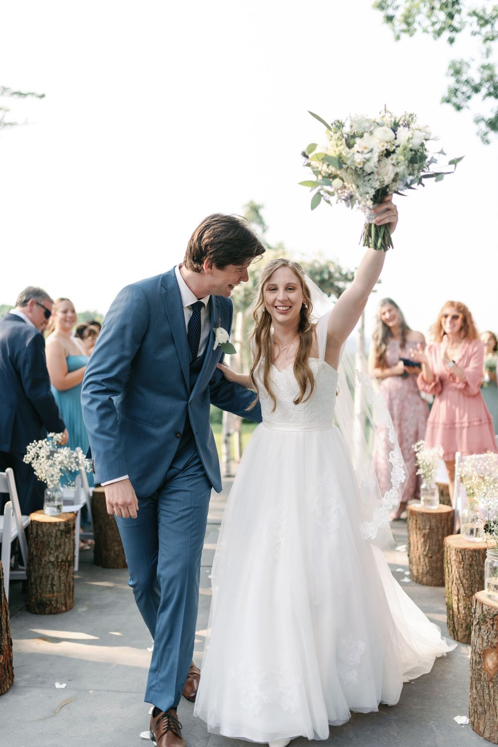 Newlyweds celebrate with joy as bride raises bouquet while walking past hay bale seating.