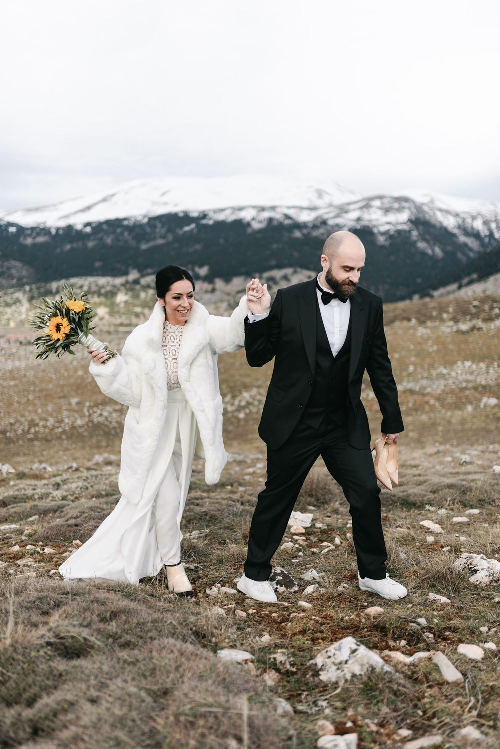 A couple in wedding attire walking together on a mountainside with snowy peaks and yellow flowers in the background.