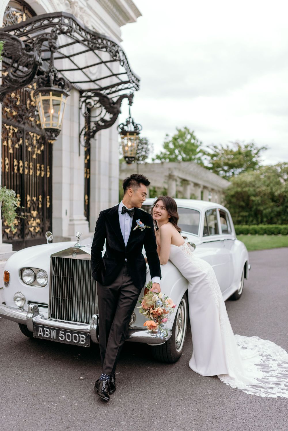A couple poses with a vintage white Rolls-Royce outside an elegant venue with ornate gates and lanterns.