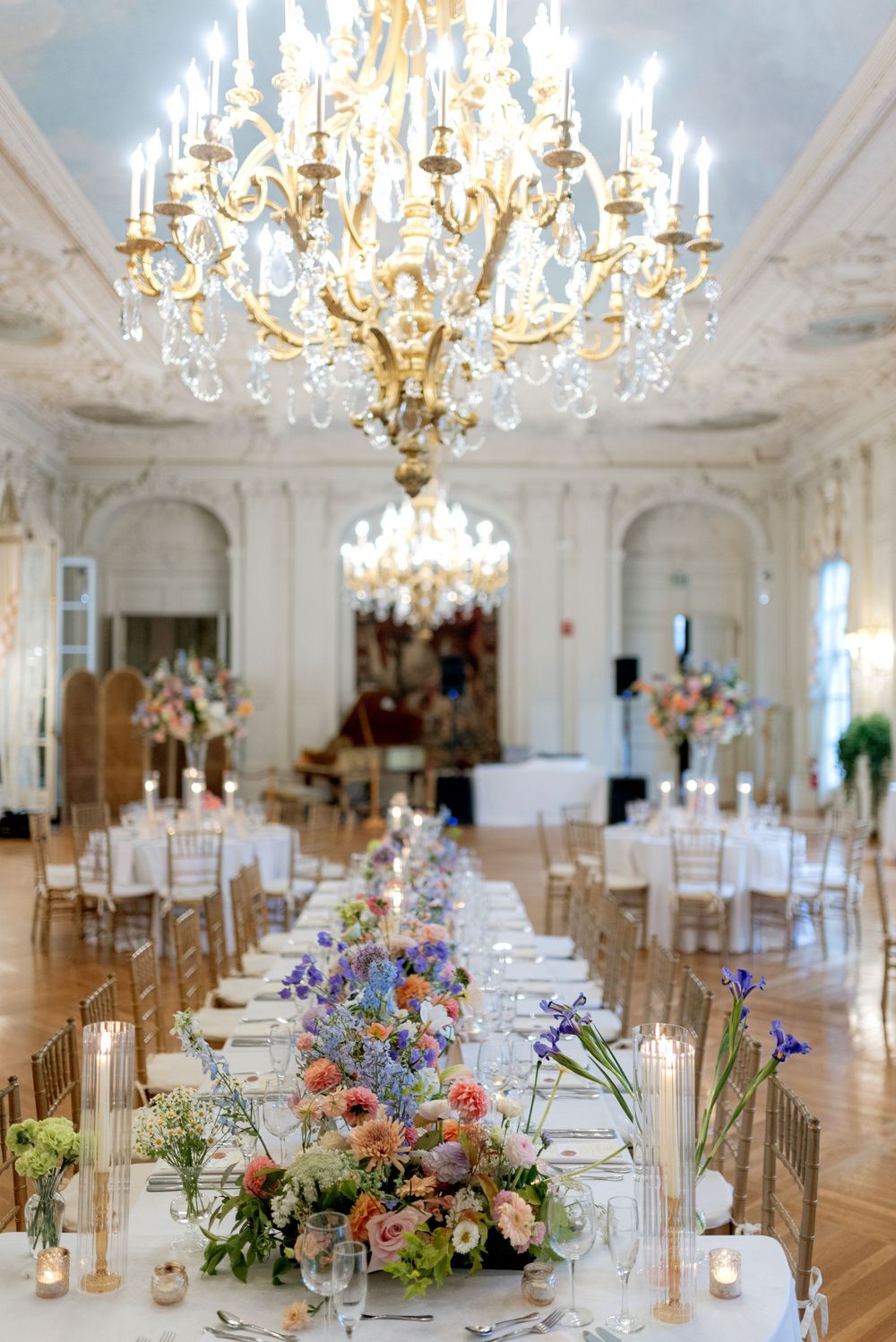 Elegant wedding reception setup in grand ballroom with crystal chandeliers and floral centerpieces on long dining tables.
