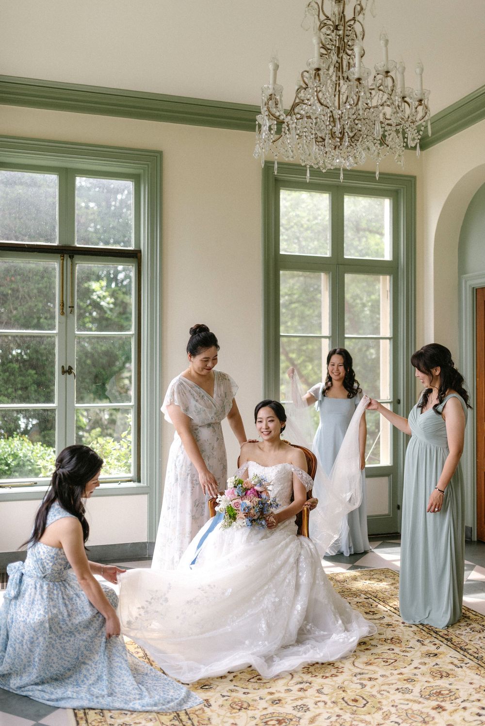 Bride in white gown holds bouquet surrounded by bridesmaids in blue dresses in elegant room with chandelier.