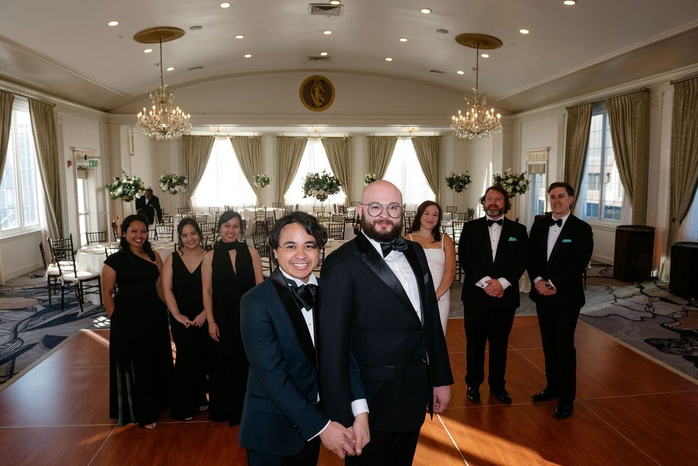 Wedding party in formal black attire poses in an elegant ballroom with chandeliers and floor-length curtains.