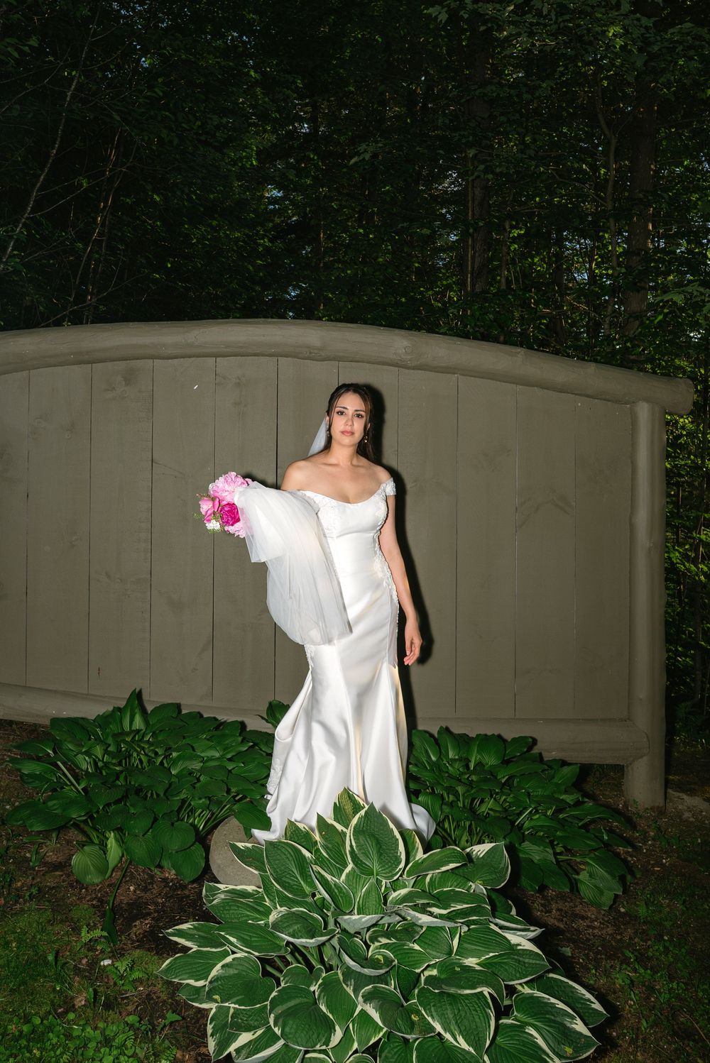 Bride in white off-shoulder mermaid gown holding pink bouquet, standing by wooden garden bridge surrounded by hosta plants.