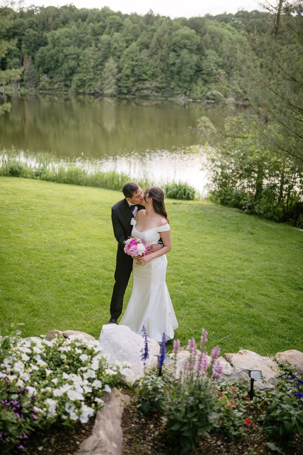 Bride and groom share a kiss on a lush green lawn beside a serene lake, surrounded by colorful garden flowers.