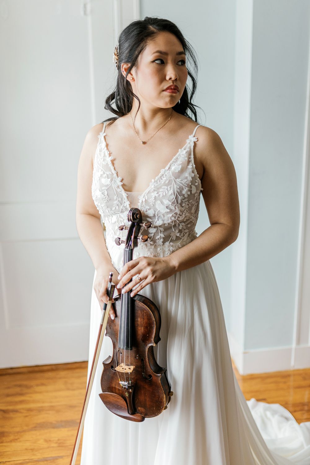 Person in white wedding dress poses with violin against neutral background.