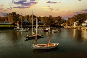 Sailboats anchored in a peaceful harbor during golden sunset with autumn trees along the shoreline.
