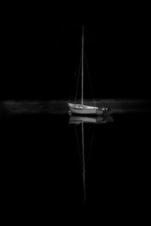 A lone sailboat reflects perfectly on calm dark waters at night, creating a mirror-like symmetrical image.
