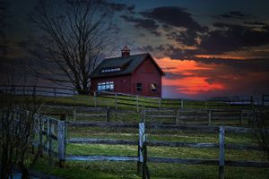 A red barn sits on a hilltop at sunset with wooden fences in the foreground and dramatic orange sky behind.
