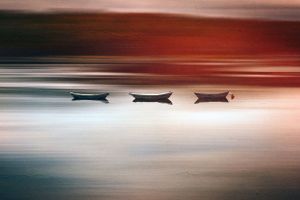 Three small boats silhouetted against calm reflective water at sunset with dramatic red and blue colored sky.