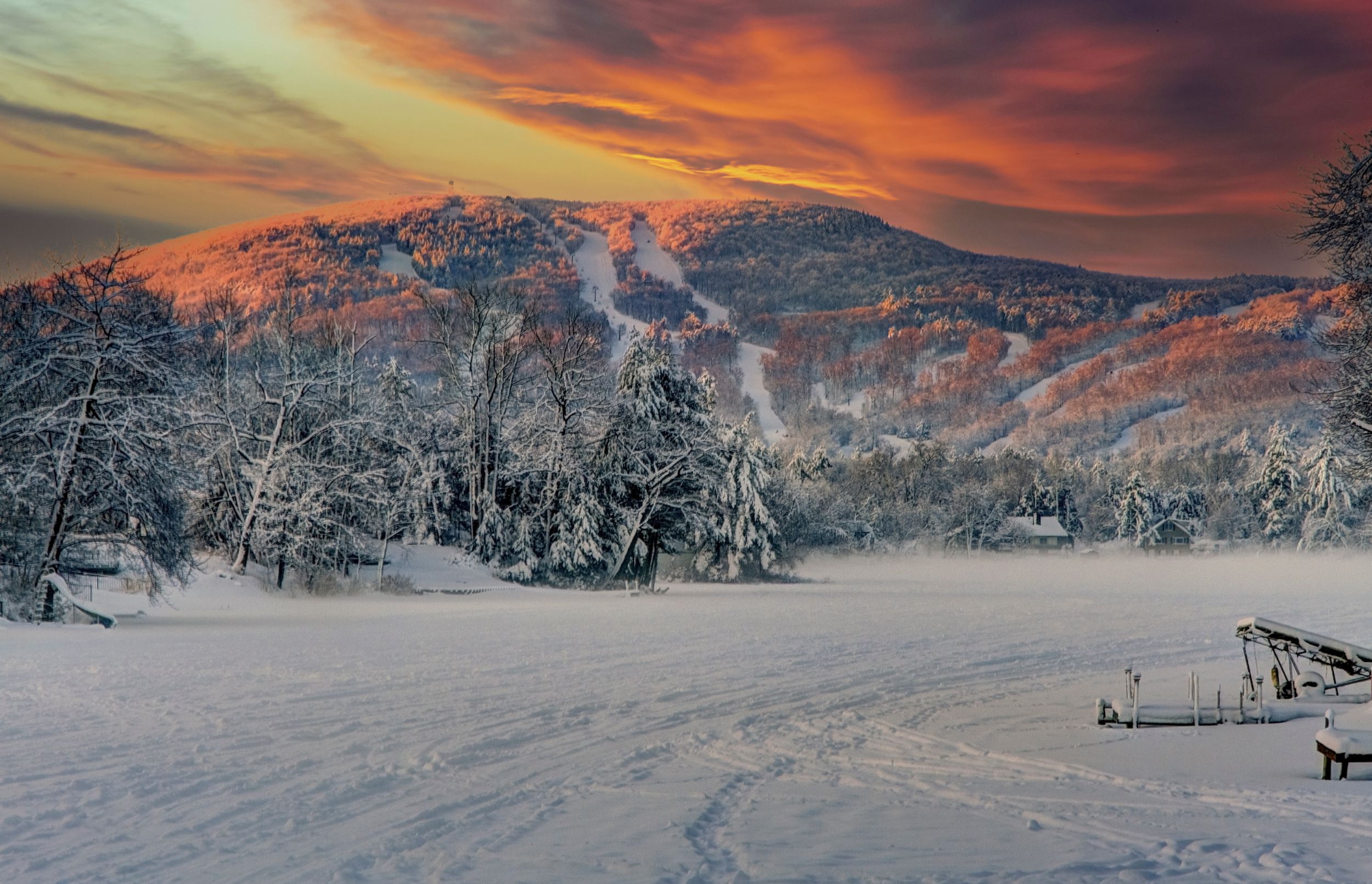 Snow-covered mountain landscape at sunset with dramatic orange sky and winding trail through pristine winter terrain.