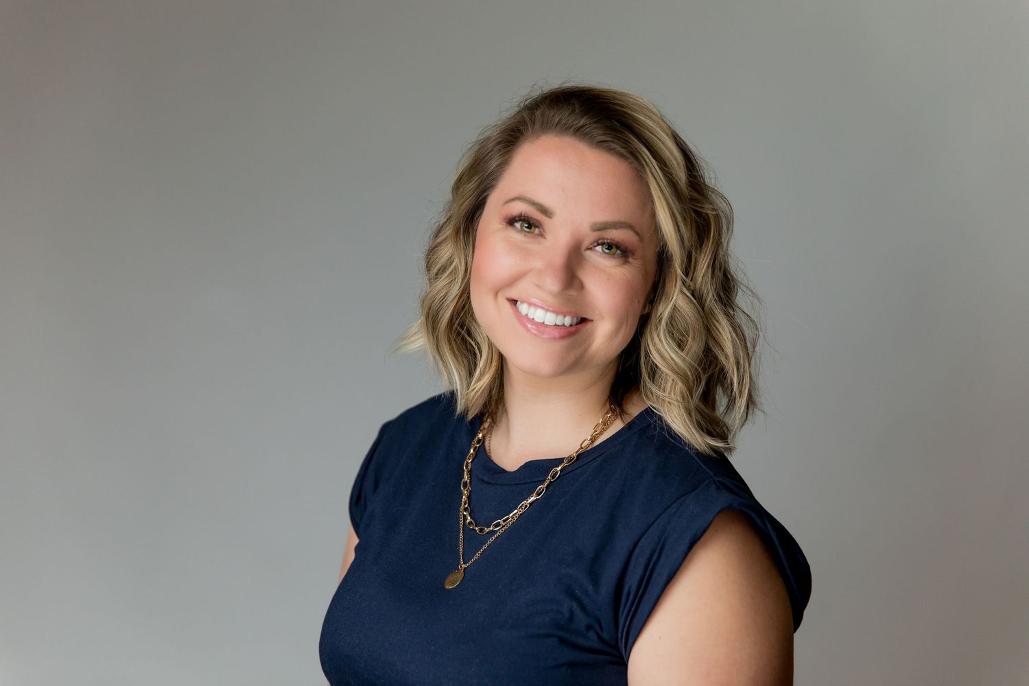 Professional headshot of a person wearing a navy blue top and necklace against a gray backdrop.