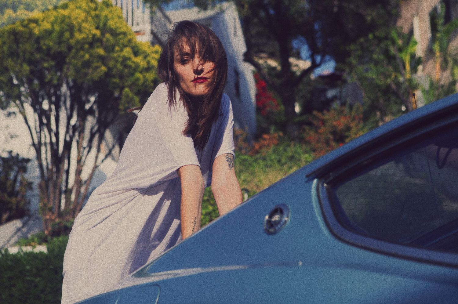 A person in casual attire poses playfully near a parked car on a sunny day.