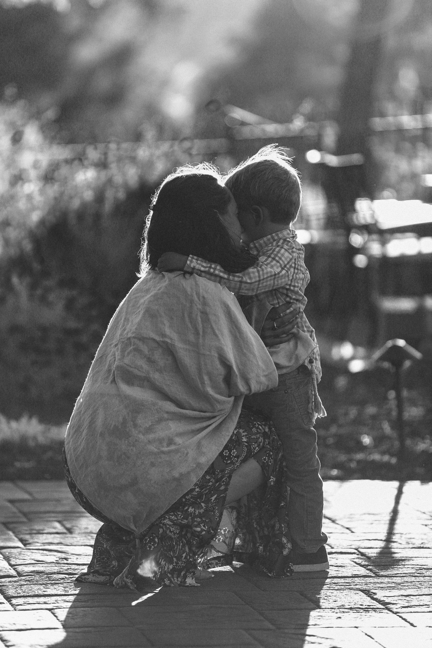 A tender black and white photograph of an adult and child sharing an intimate moment on a sunlit brick pathway.