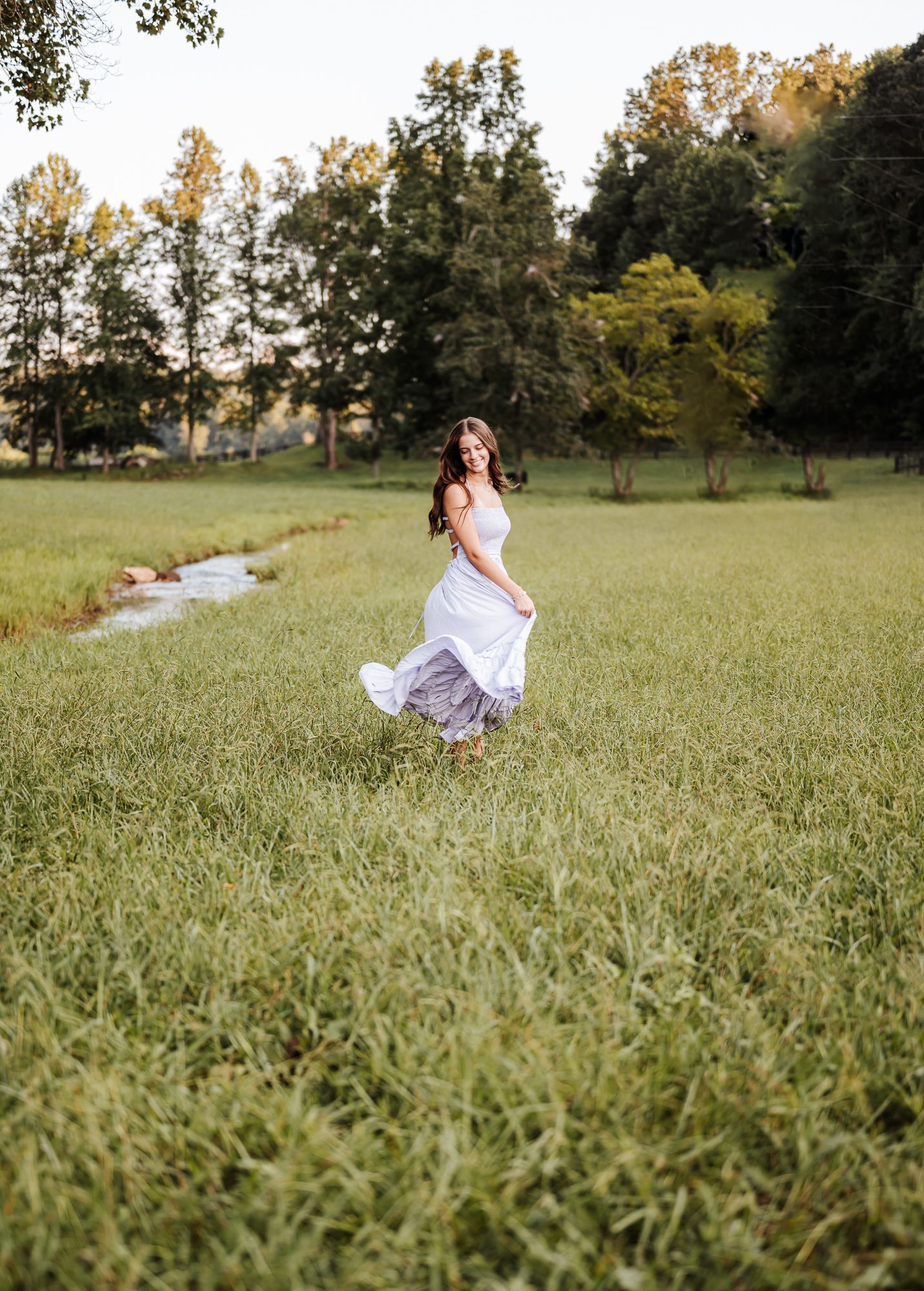 Senior in a long light blue maxi dress with flowing fabric photographed in a golden sunlit field during sunset.