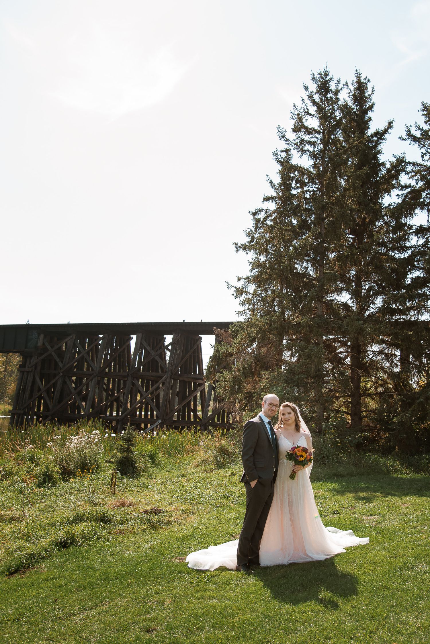 A couple stands on grassy lawn in front of rustic wooden bridge and tall evergreen trees on their wedding day.