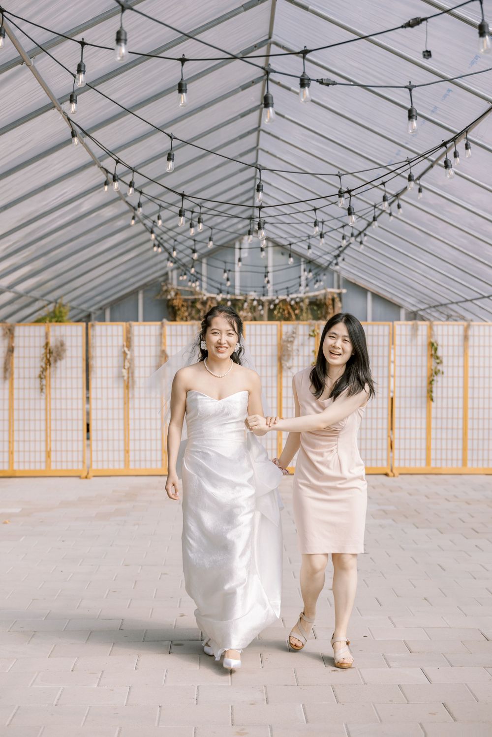 Two people in formal attire walking together under string lights in white tent venue.