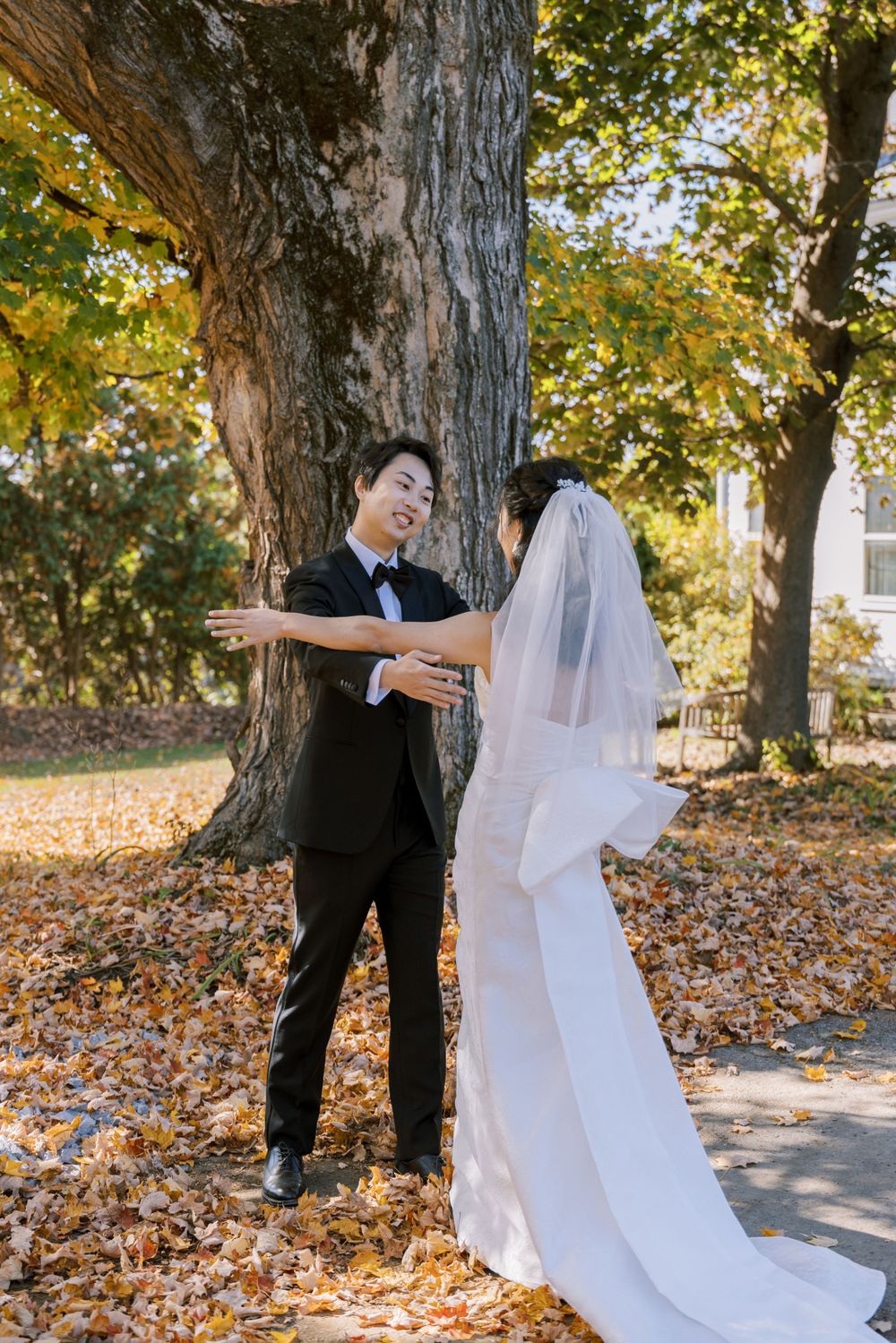 A candid wedding moment captured under a majestic tree with autumn leaves scattered on the ground.