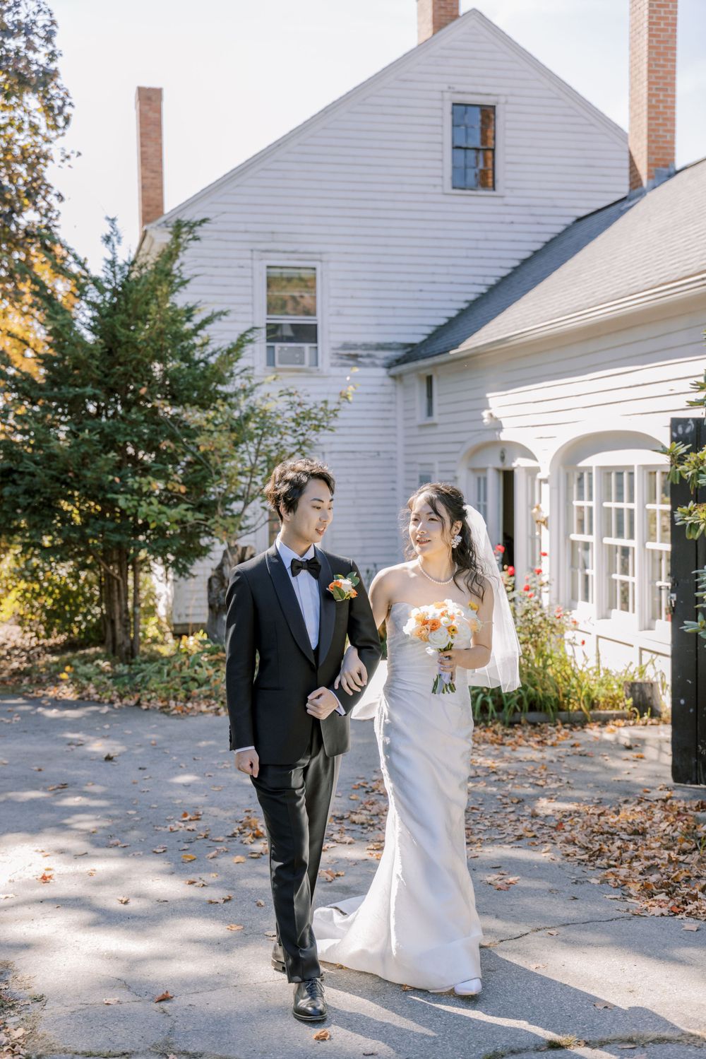 A couple walks along a pathway next to a white colonial-style house during their wedding.
