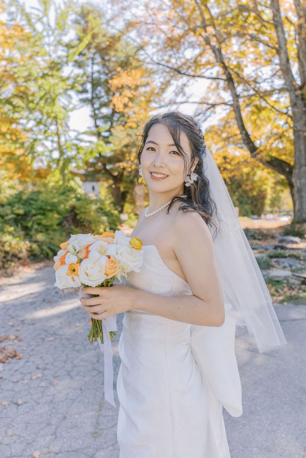 A bride in white gown holds orange and white bouquet against autumn foliage backdrop.