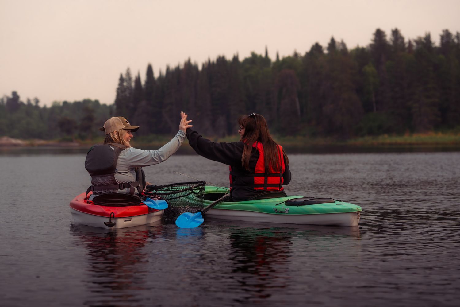 A group of kayakers paddle across a calm lake surrounded by evergreen trees at dusk.