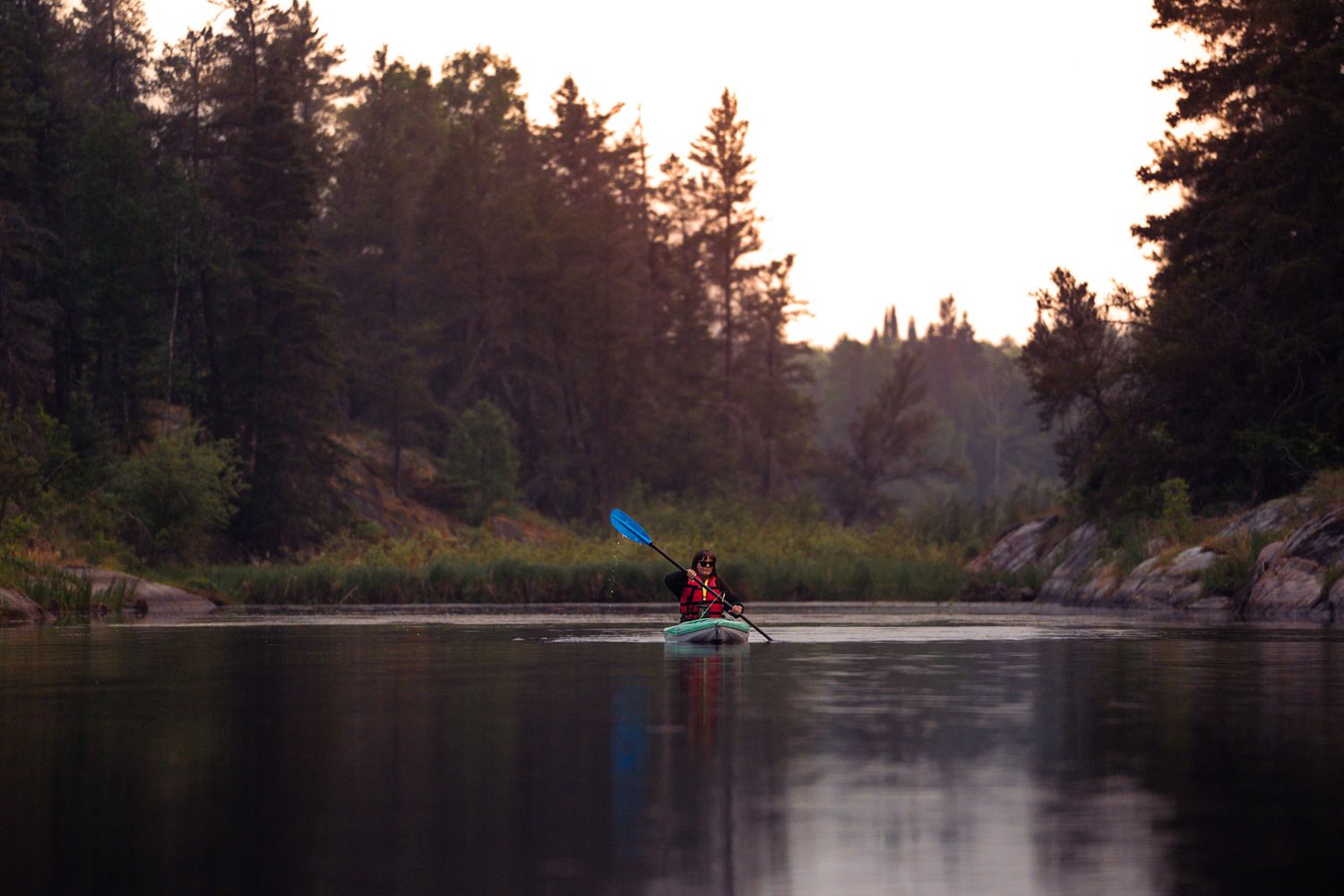 A peaceful lake scene at sunset with kayakers paddling through calm waters surrounded by pine trees.