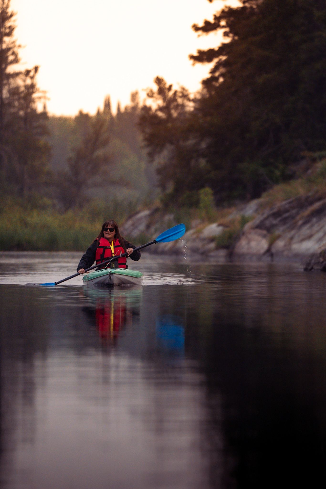 Kayaker in red jacket paddling across misty lake at sunset with blue paddle reflecting on calm waters.