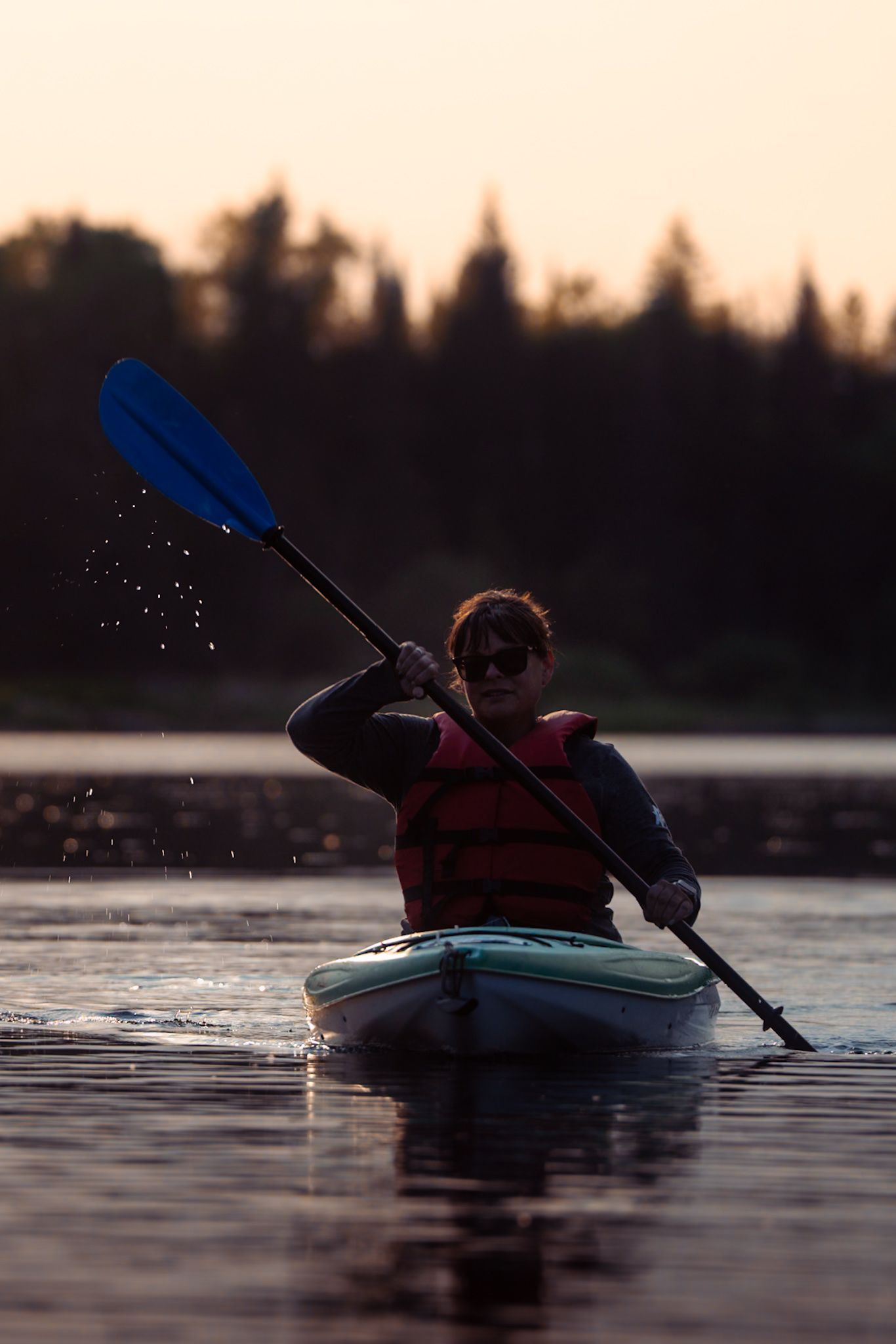 A kayaker paddles across a tranquil lake at sunset, creating ripples in the dark reflective water.