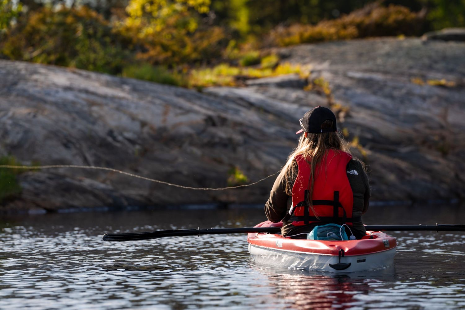 Paddler in red life vest navigating kayak near rocky shoreline.