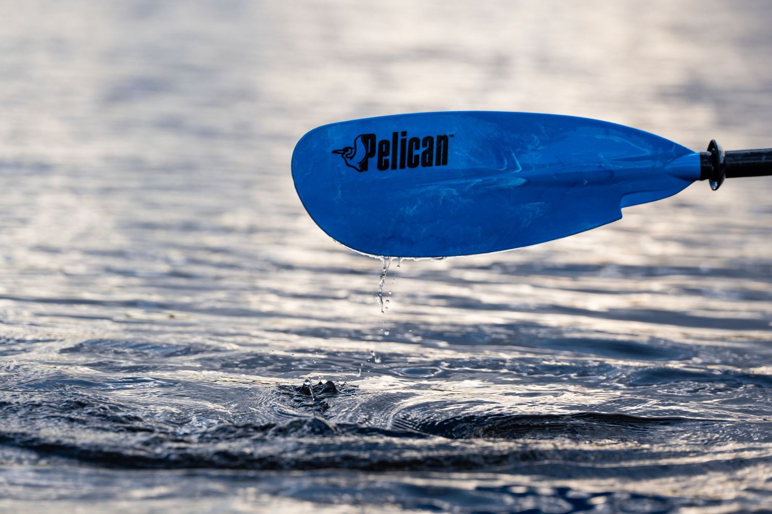 Blue Pelican kayak paddle dripping water against reflective lake surface.