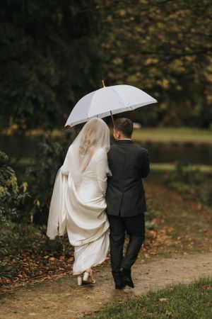 A couple walks up a leaf-covered path while holding a white umbrella.
