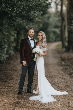 A couple in wedding attire poses on a forest path near Wootton Hall, the bride in white lace and the groom in burgundy velvet.