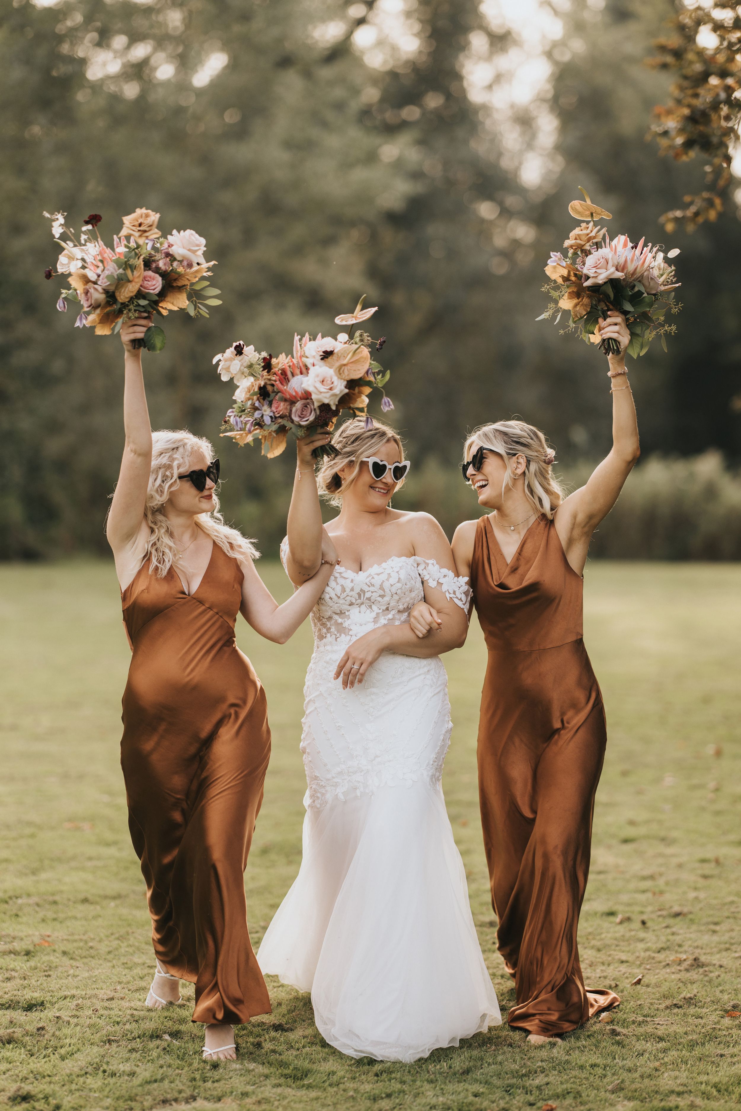 Bride in white lace gown celebrates joyfully with two bridesmaids in rust-coloured dresses holding floral bouquets outdoors.
