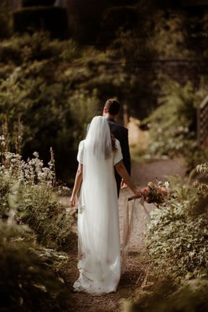 A bride in a flowing white dress and long veil walks along a garden path holding a bouquet of flowers at Hodsock Priory.