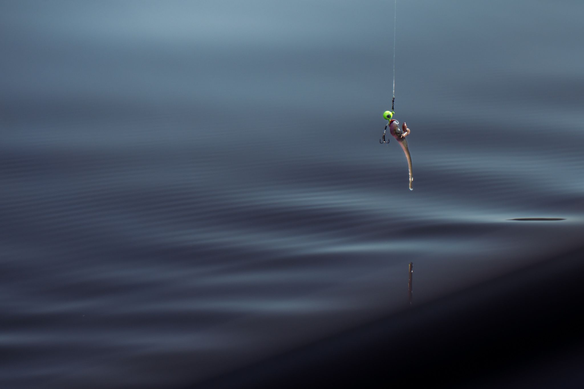 Fishing lure suspended over dark calm water with minimal ripples.