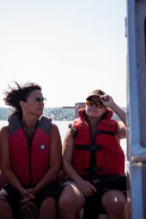 Two people in red life jackets sitting on a boat deck with wind-blown hair.
