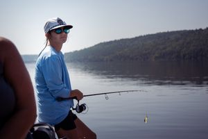 Person in blue shirt fishing from boat on calm lake with forested shore in background.