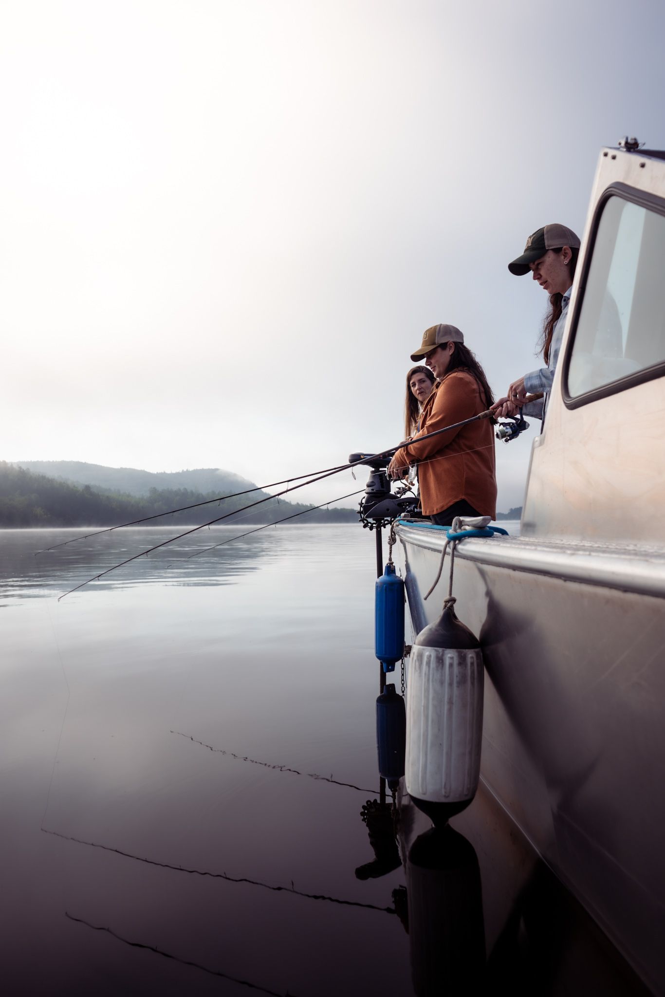 Two anglers fishing from the side of a boat on a misty morning.