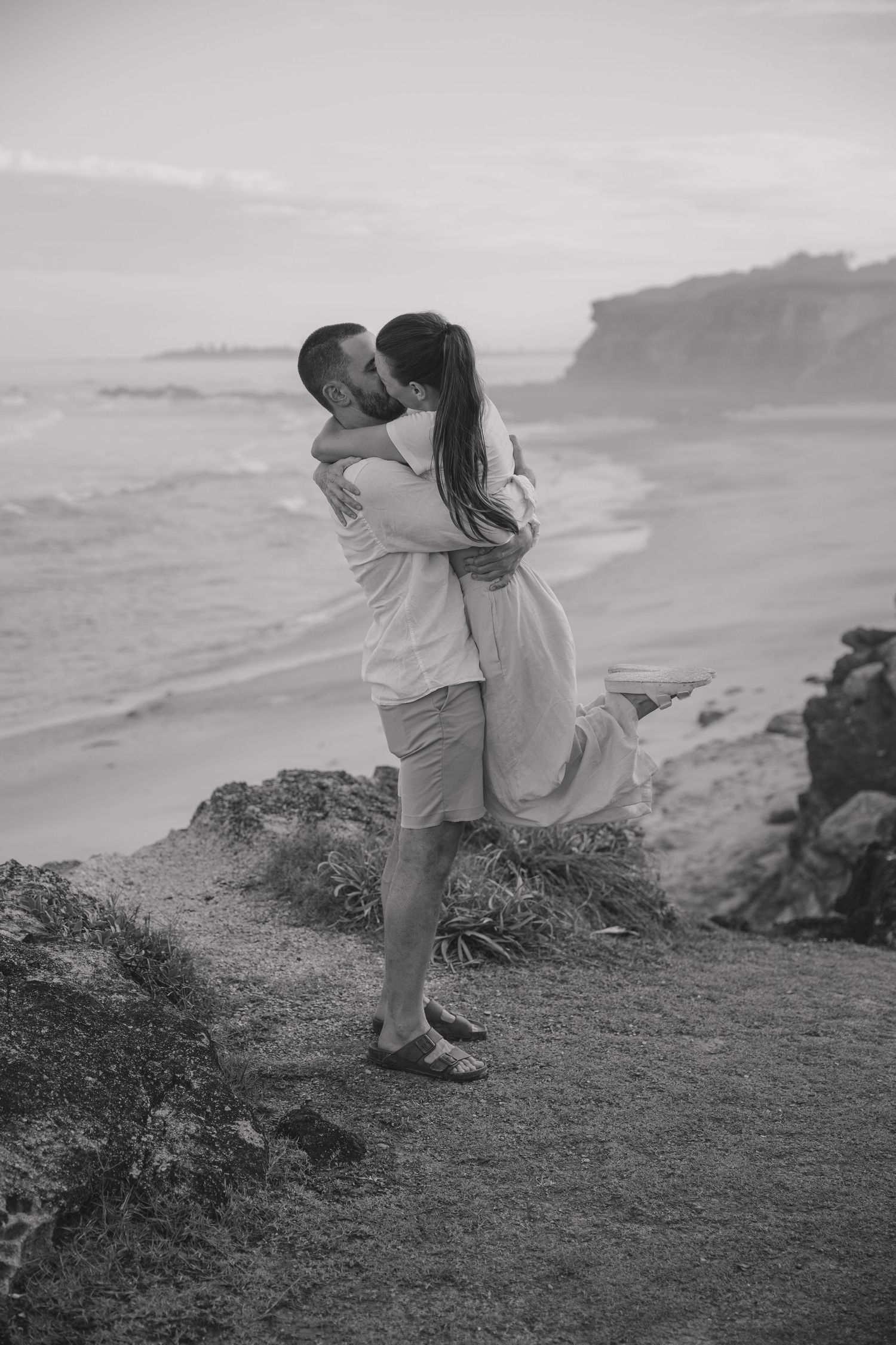 Black and white photo of a man lifting and kissing his partner on a coastal clifftop overlooking the sea.