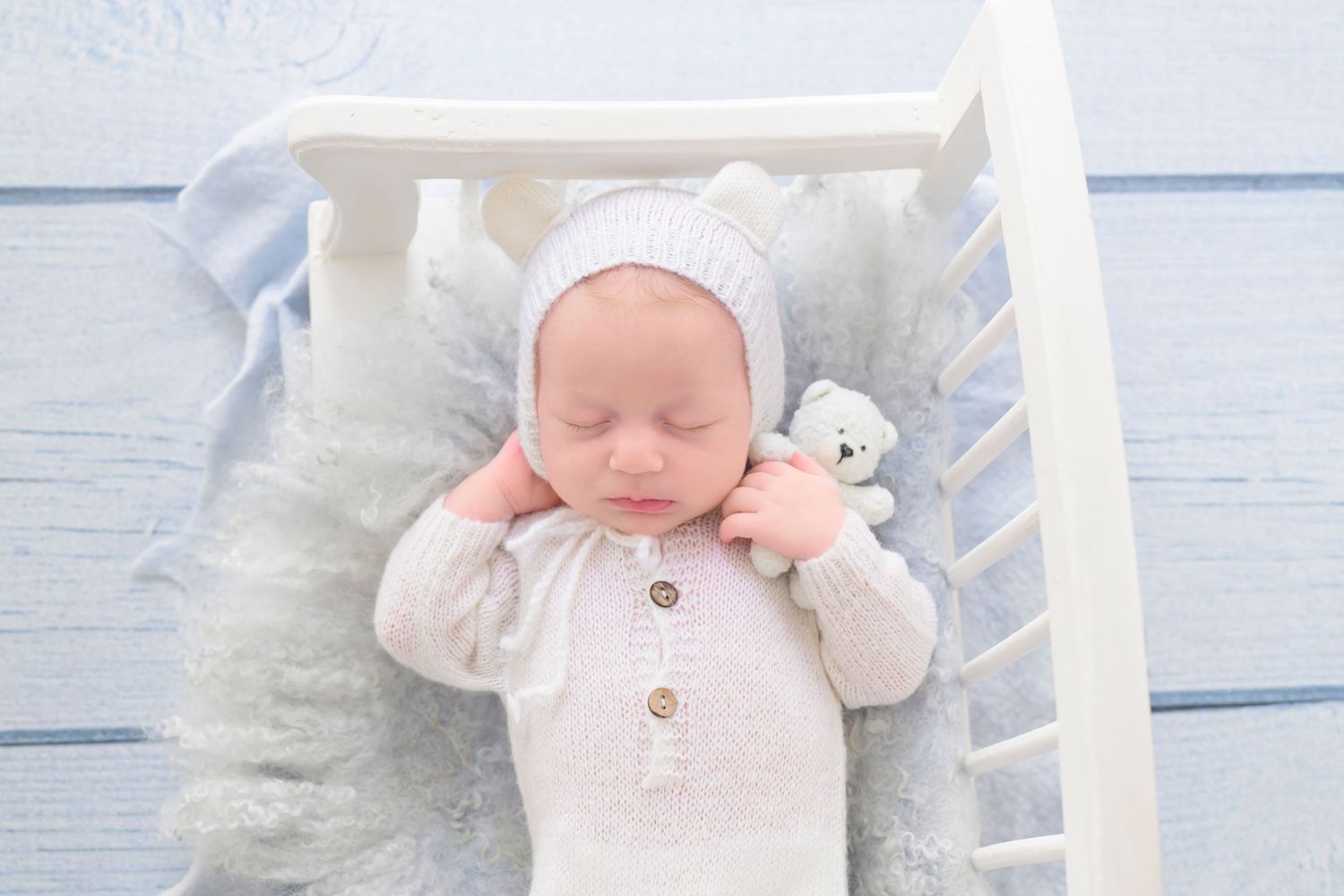 Sleeping newborn in white bear bonnet and onesie rests on blue fluffy blanket holding a small stuffed bear toy sleeping in white crib.
